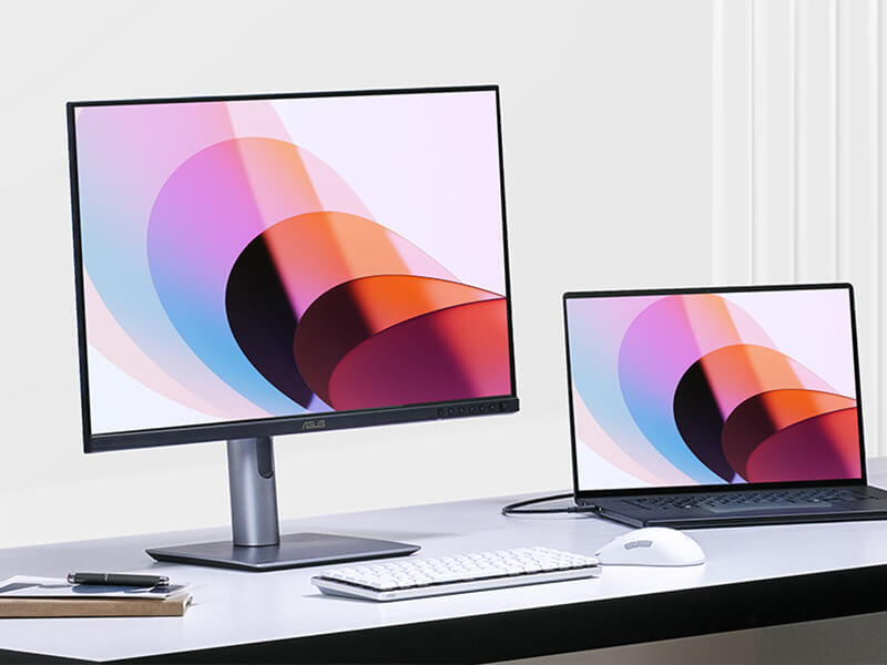 A desk setup with a ProArt monitor and a laptop, both displaying colorful abstract shapes.