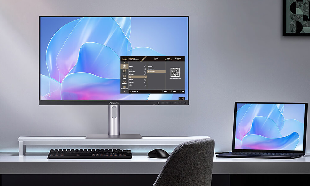 A modern desk setup with a ProArt monitor and a laptop displaying the same abstract blue and pink design. A keyboard and mouse are on the desk. The ambiance is sleek and minimalist.
